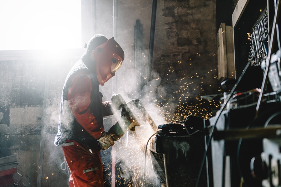 Arbeiter mit Schutzhelm und Gesichtsschutz schleift Metall mit einer Maschine, Funken und Rauch entstehen in einer industriellen Werkstatt